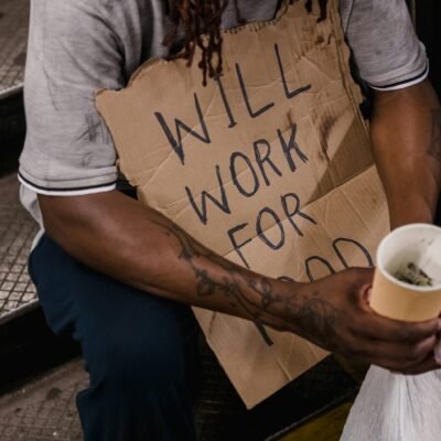 A man in need holding a sign stating 'Will Work For Food' while sitting on steps.