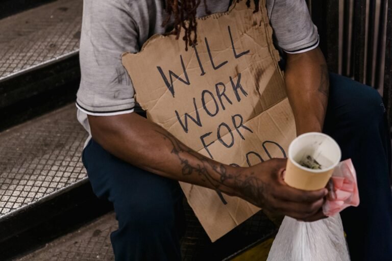 A man in need holding a sign stating 'Will Work For Food' while sitting on steps.