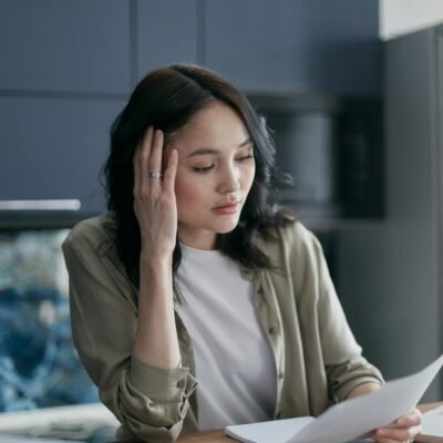 A woman looks stressed while managing bills and documents at the kitchen table.