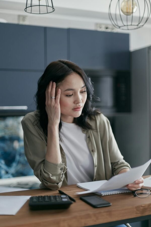 A woman looks stressed while managing bills and documents at the kitchen table.