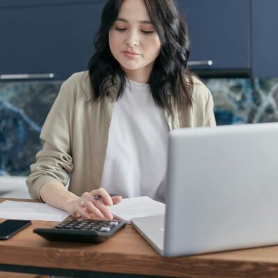 A young woman calculating finances at home using a laptop and calculator, reflecting a focused budgeting process.