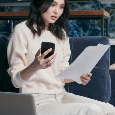 A young woman uses her smartphone and laptop to manage finances, appearing focused and concerned.