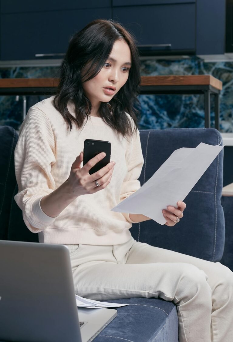 A young woman uses her smartphone and laptop to manage finances, appearing focused and concerned.